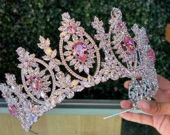 May include: A silver-toned tiara with clear and pink gemstones. The crown features a floral design with large, oval-shaped pink stones at the center and smaller pink stones throughout. The tiara is held by a hand against a green background.