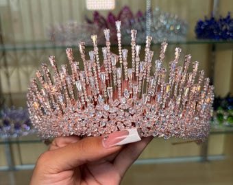 May include: A rose gold tiara with a sparkling design. The crown features numerous clear crystals and vertical accents, creating a dazzling effect. The tiara is designed for special occasions.