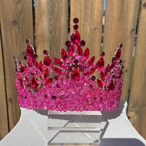May include: A vibrant pink and red jeweled crown. The crown features large, teardrop-shaped red gems and smaller, round red gems. The base is a solid band of pink beads, and the crown is displayed on a clear stand.