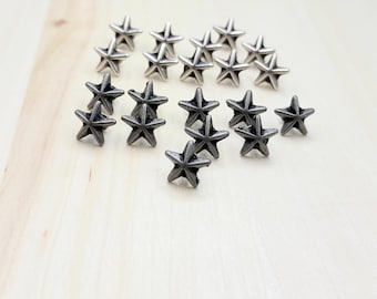 Puede incluir: Colección de pequeños tachuelas metálicas plateadas en forma de estrella. Las tachuelas están dispuestas sobre una superficie de madera clara. Cada estrella tiene cinco puntas y un aspecto ligeramente texturizado.