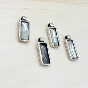 May include: Four silver pendant charms with rectangular clear glass stones. The charms are on a light wood surface.