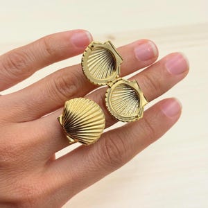 May include: Two gold-colored seashell-shaped rings. One ring is open, revealing the interior, while the other is closed. The rings have a textured, ridged design, and are set against a neutral background. The rings are likely jewelry.