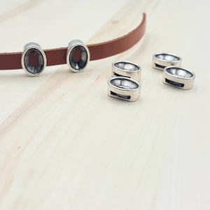 May include: Five silver oval-shaped metal beads with a hole in the center. The beads are on a light wood surface. Two of the beads are on a brown leather strap.
