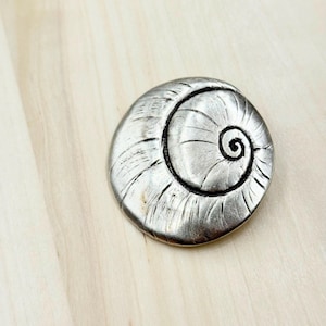 May include: A silver-coloured, spiral-shaped decorative object. The object has a textured surface with a dark, spiralling centre. The design resembles a seashell. The object is circular and sits on a light-coloured wooden surface.