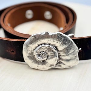 May include: A brown leather belt with a silver, spiral-shaped buckle. The buckle is a detailed, textured design.