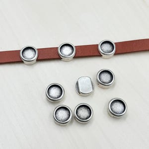 May include: Silver-toned metal round bezel settings for jewelry making. The bezels are shown on a brown leather strap and on a white surface.