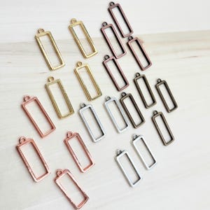 May include: Assortment of 15 rectangular metal pendant frames in various finishes, including silver, gold, copper, and bronze. The frames are all the same size and shape, and they have a simple, elegant design.