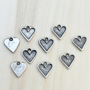 May include: A collection of small, silver-toned heart charms. Each charm has a loop at the top for stringing and a slightly textured surface. The hearts are arranged on a light-colored wooden surface.