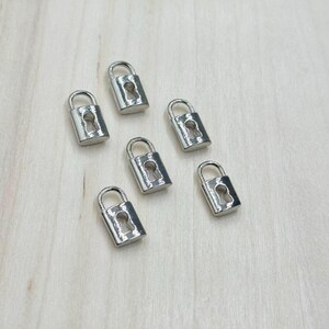 May include: Six small, silver-colored padlocks with keyhole cutouts are arranged on a light-colored wooden surface. Each padlock has a curved handle. The padlocks are evenly spaced and appear to be new.