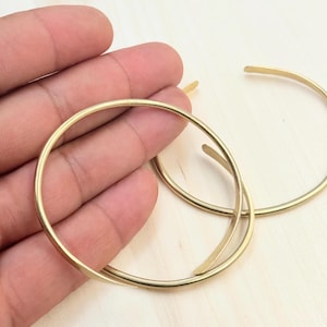 May include: Two gold-colored hoop earrings. One earring is partially overlapping the other. The earrings are held in a hand against a light-colored wooden surface. The earrings have a simple, minimalist design.