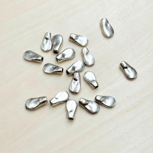 May include: A collection of silver-colored, teardrop-shaped metal beads scattered on a light wood surface. Each bead has a textured, slightly irregular surface and a small hole at the narrow end, suggesting they are for jewelry making or crafts.