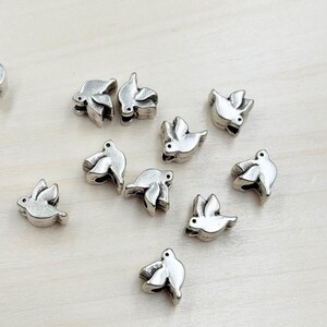 May include: A group of silver dove-shaped beads, scattered on a light wood surface. The beads are small and have a delicate design.