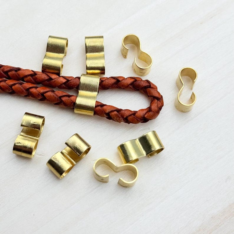 Op de afbeelding: Een verzameling van goudkleurige metalen sluitingen voor lederen koord of band. De sluitingen zijn in verschillende vormen, waaronder een eenvoudige lus en een figuur acht.