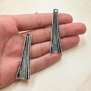 以下が含まれることがあります： 銀色の細長いペンダントチャームが2つ。テクスチャ加工されたリブのデザインです。各チャームの上部には、紐を通すための小さな穴があります。チャームはニュートラルな背景に表示されています。