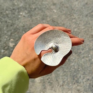 May include: A large, silver-toned ring with a textured, fan-like design. The ring features a central, rounded element and a unique, open design. The ring is worn on a hand with a light green sleeve in the background.