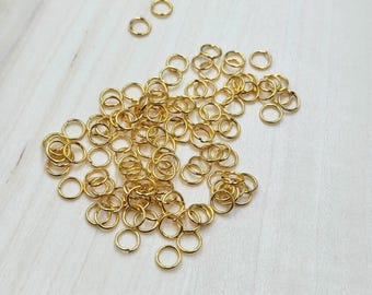 May include: A collection of small, gold-colored jump rings scattered on a light-colored wooden surface. The rings are circular and appear to be made of metal, likely for jewelry making or crafting. The rings vary slightly in size.