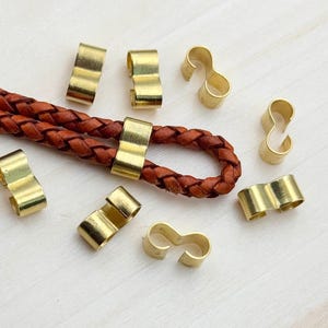 Op de afbeelding: Een close-up van goudkleurige metalen sluitingen voor lederen koord. De sluitingen zijn in verschillende vormen, waaronder een eenvoudige lus en een figuur acht. De sluitingen zijn gerangschikt op een licht houten oppervlak.