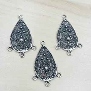 May include: Three silver-toned teardrop-shaped pendants with intricate embossed designs. Each pendant features a central starburst pattern, surrounded by smaller circular and linear details. The pendants have loops for attachment and are displayed on a light wooden surface.
