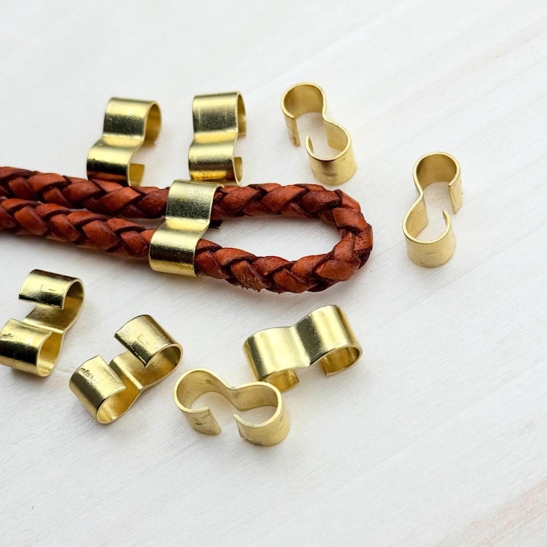Op de afbeelding: Een close-up van acht gouden metalen sluitingen voor leren koord of touw. De sluitingen zijn S-vormig en hebben een gladde, glanzende afwerking.