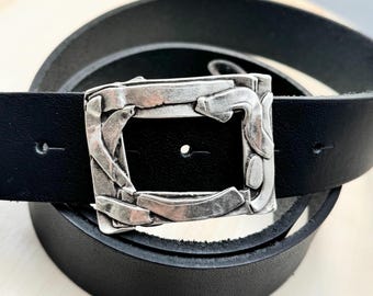 May include: A black leather belt with a silver, textured buckle. The buckle has a unique, abstract design.