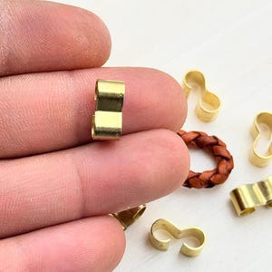Op de afbeelding: Een close-up van een gouden metalen sluiting, met een gevlochten leren koord op de achtergrond. De sluiting is ontworpen om twee stukken koord of touw aan elkaar te verbinden.