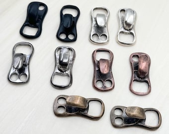 May include: A collection of metal zipper pulls in various finishes, including black, silver, and bronze. Each pull features a distinctive design with a loop and two holes for attachment, suitable for a variety of sewing and crafting projects.