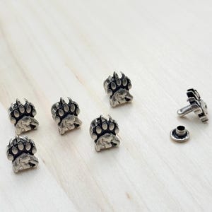 May include: Six silver bear paw print metal studs with one silver rivet. The studs have a raised design of a bear paw print.