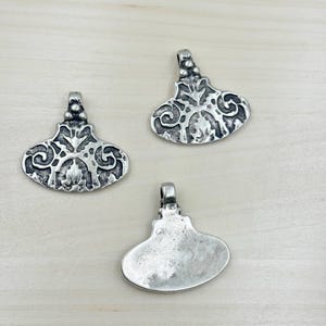 Puede incluir: Tres colgantes plateados con un diseño ornamentado y simétrico. Cada colgante tiene un lazo en la parte superior para una cadena y un patrón detallado de espirales y formas. Los colgantes se muestran sobre una superficie de madera clara.