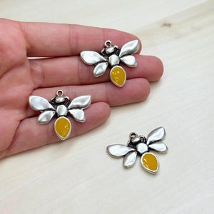 May include: Three silver-toned bee charms with yellow enamel accents. Each charm features detailed wings, a body, and a loop for attaching to jewelry or crafts. The bee charms are a charming addition to any project.