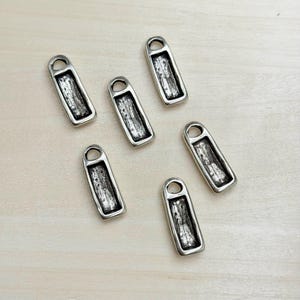 May include: Six silver-toned metal zipper pulls with a rectangular shape and a textured, weathered appearance. Each pull has a small loop at the top. The pulls are scattered on a light wood surface.