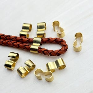 Op de afbeelding: Een close-up van acht gouden metalen koord eindkappen. De koord eindkappen zijn ontworpen om een ​​leren koord op zijn plaats te houden. De koord eindkappen zijn willekeurig gerangschikt op een wit houten oppervlak.