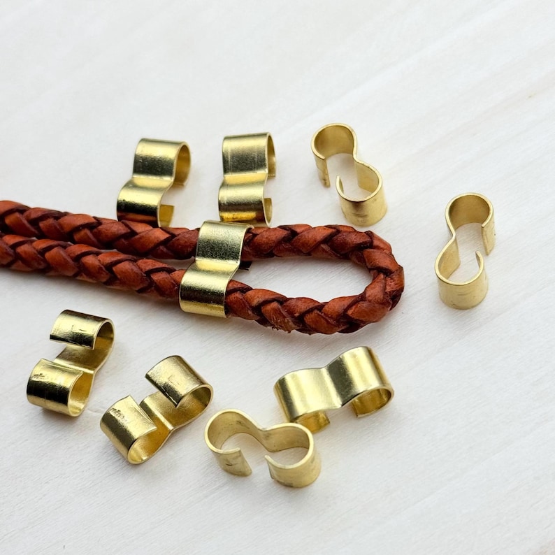 Op de afbeelding: Een close-up van acht gouden metalen koord eindkappen. De koord eindkappen zijn ontworpen om een ​​leren koord op zijn plaats te houden. De koord eindkappen zijn willekeurig gerangschikt op een wit houten oppervlak.