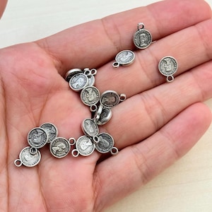 以下が含まれることがあります： ジュエリー作りに使用する、ループ付きの小さな銀色のコインチャーム。各チャームには人物の横顔と「SECOND ELIZABETH QUEEN」の文字が描かれています。チャームは明るい背景に散らばっています。