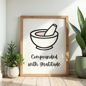 May include: A framed print featuring a black and white illustration of a mortar and pestle. The text "Compounded with gratitude" is below the illustration. The frame is made of light brown wood.