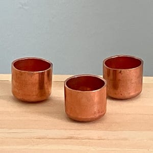May include: Three small, cylindrical copper cups with a polished finish. The cups are arranged on a light-colored wooden surface against a pale blue background. The open tops of the cups reveal a reddish-brown interior.