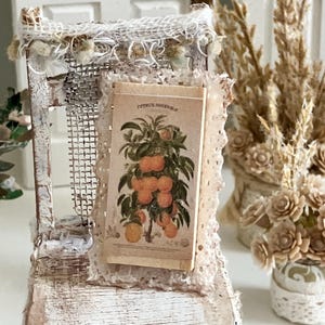 May include: A miniature, distressed white wooden stand with a vintage botanical print featuring oranges. The stand is adorned with lace and mesh details, creating a rustic, handcrafted aesthetic. Dried floral arrangements are visible in the background.