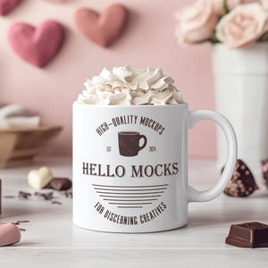 K&ouml;nnte beinhalten: Eine wei&szlig;e Keramiktasse mit einem schwarz-wei&szlig;en Design, auf dem "High-Quality Mockups Est. 2014 Hello Mocks For Discerning Creatives" steht. Die Tasse ist mit Schlagsahne gef&uuml;llt und steht auf einer wei&szlig;en Oberfl&auml;che.