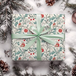 May include: A white gift box with a green ribbon and a floral pattern featuring birds and red berries. The box is surrounded by snow and pine branches.