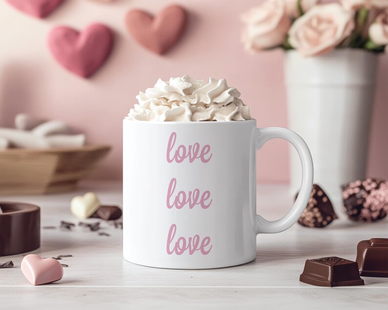 K&ouml;nnte beinhalten: Eine wei&szlig;e Keramiktasse mit dem dreimal wiederholten Wort 'love' in Rosa. Die Tasse ist mit Schlagsahne gef&uuml;llt und steht auf einer wei&szlig;en Oberfl&auml;che mit Schokoladenbonbons.
