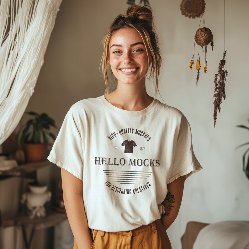 May include: A woman wearing a white t-shirt with the text "HIGH-QUALITY MOCKUPS EST 2024 HELLO MOCKS FOR DISCERNING CREATIVES" printed on it. She is standing in front of a white wall with a macrame hanging behind her.