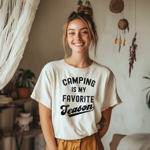 May include: A woman wearing a white t-shirt with the text "Camping is my favorite season" in black lettering. The text is in a retro font style.
