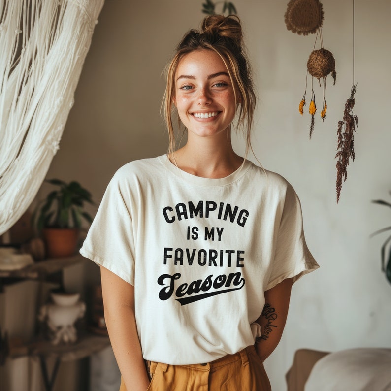 May include: A woman wearing a white t-shirt with the text "Camping is my favorite season" in black lettering. The text is in a retro font style.