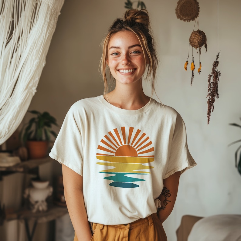 May include: A white t-shirt with a retro sunset graphic in shades of orange, yellow, and blue. The graphic features a sun with rays and a wavy line representing water.