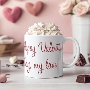 K&ouml;nnte beinhalten: Eine wei&szlig;e Keramiktasse mit einer roten Aufschrift, die "Happy Valentine's Day, my love!" lautet. Die Tasse ist mit Schlagsahne gef&uuml;llt und steht auf einer wei&szlig;en Oberfl&auml;che.