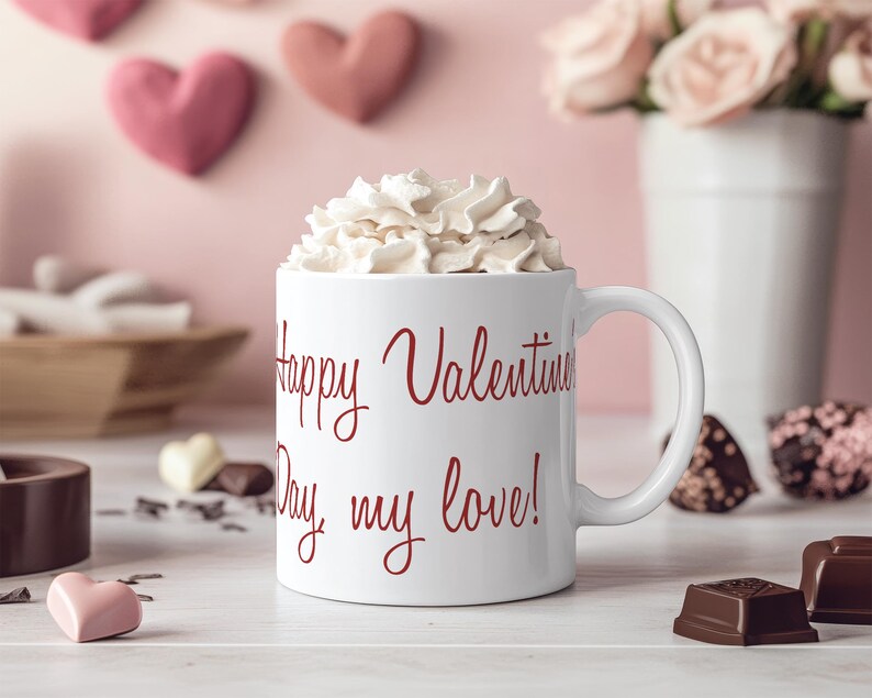 K&ouml;nnte beinhalten: Eine wei&szlig;e Keramiktasse mit einer roten Aufschrift, die "Happy Valentine's Day, my love!" lautet. Die Tasse ist mit Schlagsahne gef&uuml;llt und steht auf einer wei&szlig;en Oberfl&auml;che.