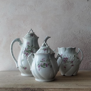 Peut inclure: Un service à thé en porcelaine blanche composé d'une théière haute avec couvercle, d'un sucrier couvert et d'un pot à lait. Chaque pièce présente un motif floral délicat peint à la main avec de petites fleurs roses et des feuilles vertes.
