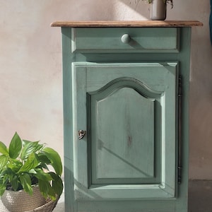 Peut inclure: Un meuble en bois peint en vert sauge doté d'un tiroir unique avec un petit bouton rond et d'une porte de placard inférieure avec un panneau incurvé. Le meuble présente un plateau en bois naturel et une charnière en métal foncé sur le côté.