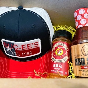 May include: A gift set featuring a black and red trucker hat with a white mesh back and a patch that reads "BUC-EE'S EST. 1982". Also included are a jar of Texas Round Up BBQ Rub and a bottle of Original BBQ Sauce.