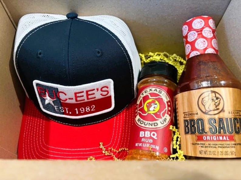May include: A gift set featuring a black and red trucker hat with a white mesh back and a patch that reads "BUC-EE'S EST. 1982". Also included are a jar of Texas Round Up BBQ Rub and a bottle of Original BBQ Sauce.