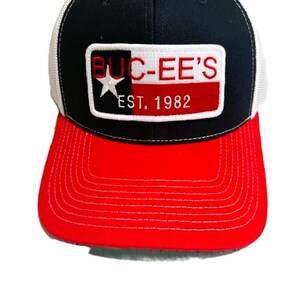 May include: A black, red, and white baseball cap with a mesh back. The front features a rectangular patch with the text "BUC-EE'S" in red letters, a white star, and the text "EST. 1982". The brim is red.